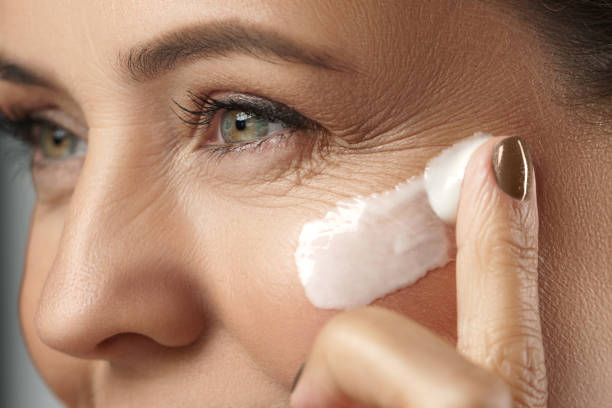 Beautiful middle aged woman applying anti aging cream on her face on gray